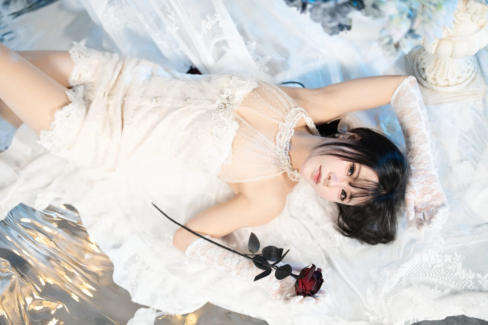 Figure in a white lace dress lying among fabric, holding a dark rose
